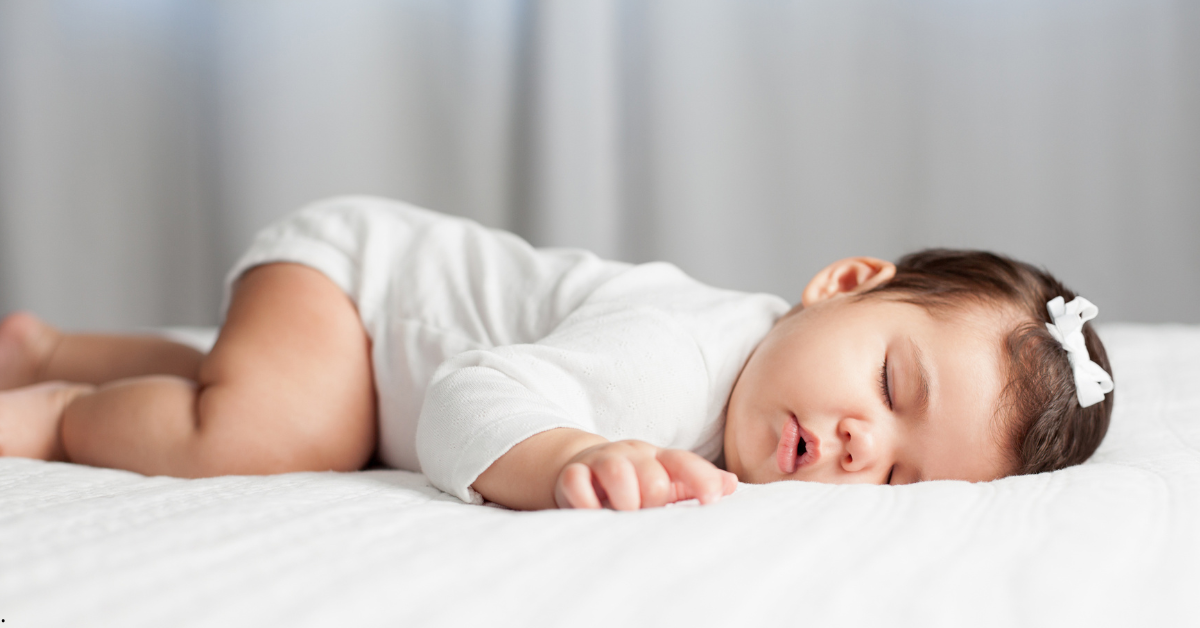 Bebê dormindo de lado com roupa branca e laço no cabelo, em sono tranquilo