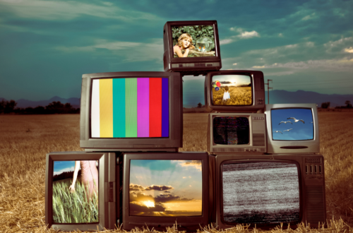 TVs antigas empilhadas em um campo seco, exibindo imagens coloridas e estáticas