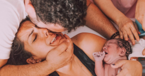 Mulher emocionada sendo amparada logo após o parto com o bebê recém-nascido no colo